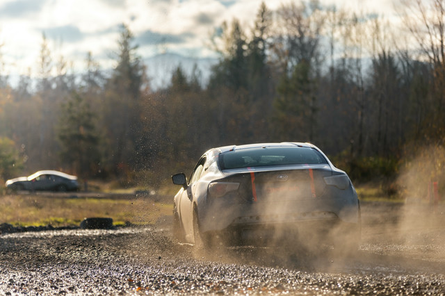 A car begins to initiate a sliding turn to the right. Another car has exited on the next straight over and is pushing ahead. The cars are black coupes with orange Dirtfish accents.