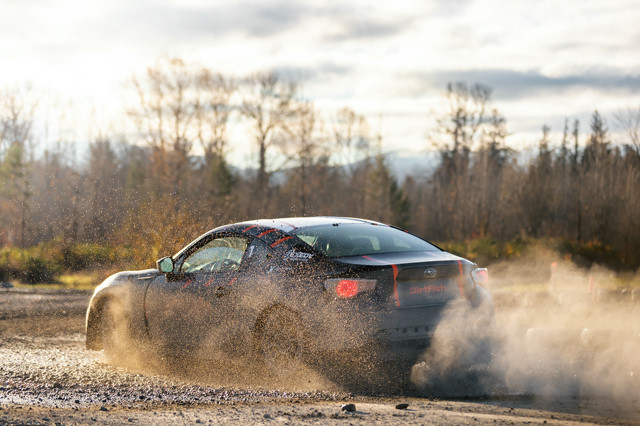 A car kicks up dirt and smoke as it turns in the dirt. The car is a black coupe with orange highlights.