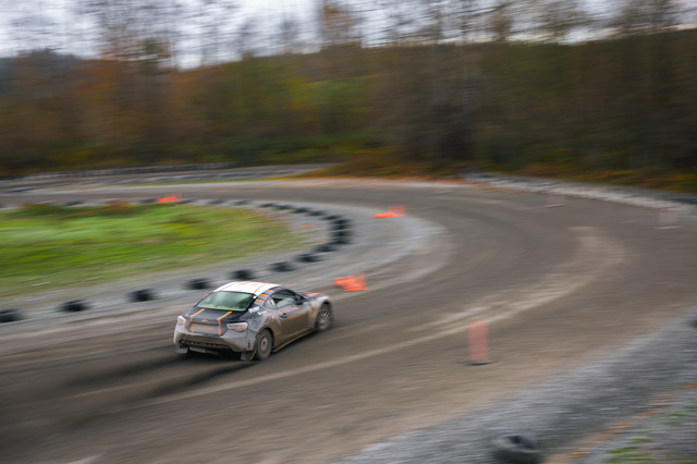 A black coupe with orange highlights begins to initiate a left hand sliding turn through a long flowing hairpin turn. Orange cones mark the course edges. A forest is off to the right.