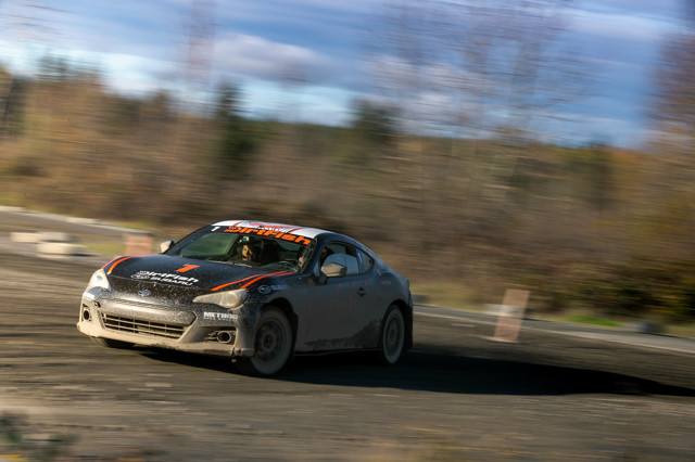 A black car with orange highlights slides out of a turn. The co-driver is reading notes aloud.