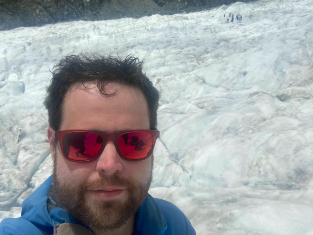 Me talking a self-photo on top of a glacier in New Zealand. A group of other hikers can be seen in the distance. Otherwise, it's just ice.