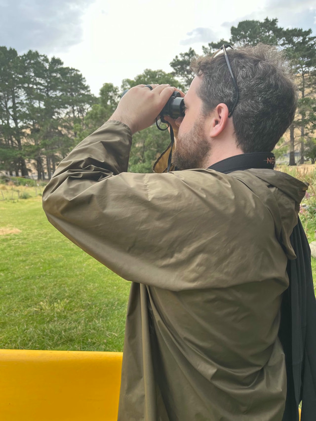 A bearded man with dark hair in a camo jacket looks through a pain of binoculars.