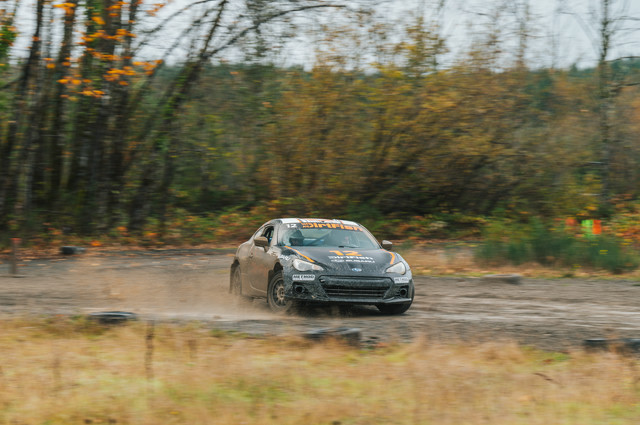 A black and orange car sliding through a forest turn.