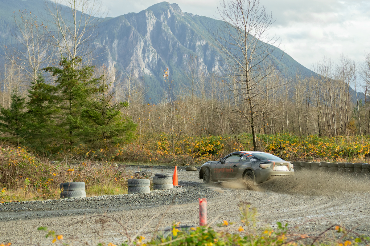 A black and orange sports car making a tight turn in gravel. Tires and orange cones mark the course layout, a mountain looms in the background.