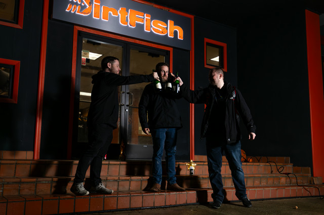 Three men stand on a set of steps, cheering to each other with champagne. The orange Dirtfish text glows in the background.