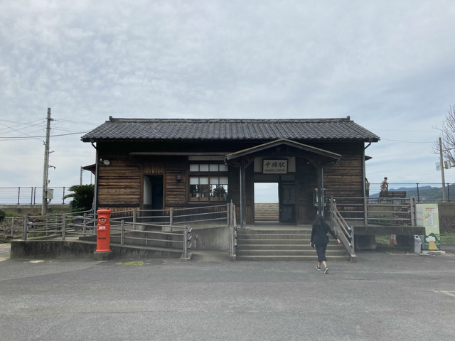 A small wooden train station on the water. A woman is walking into it. She is wearing all black.