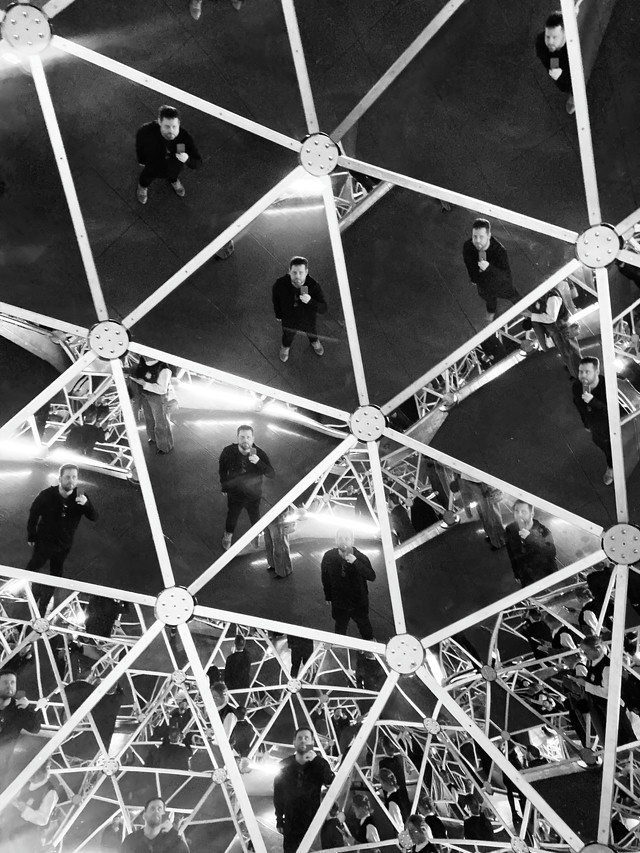 A man dressed in black takes a photo of himself in a kaleidoscope of triangle shaped mirrors. The photo is altered to be black and white.