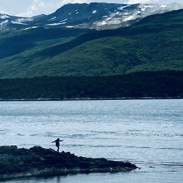 A silhouette of a woman walks on rocks on a small islet. Mountains loom in the background.