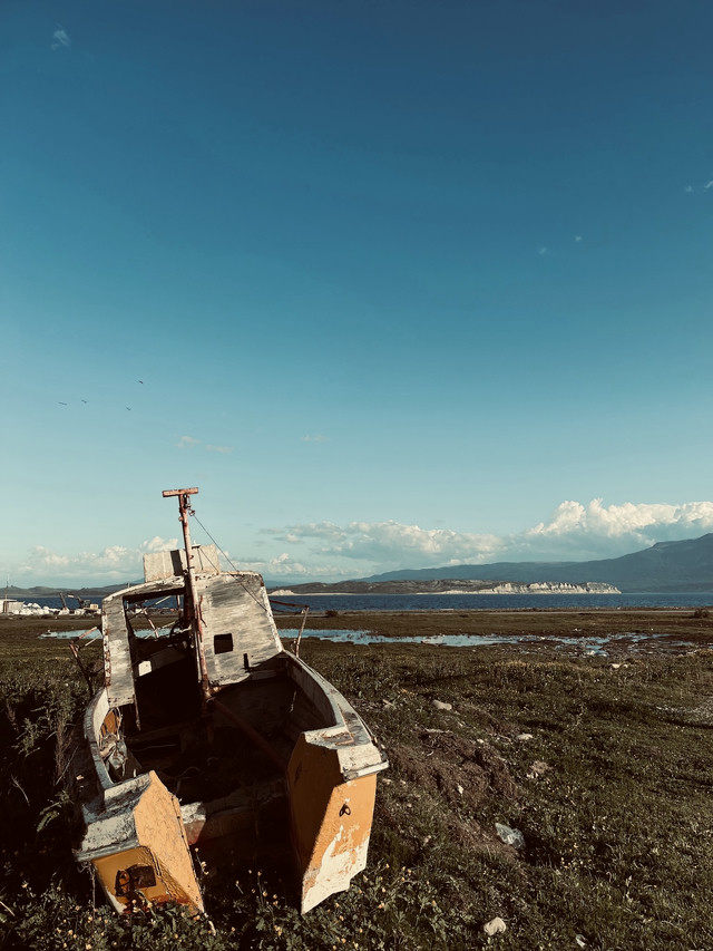 A broken down yellow boat, beached in a field. Water and mountains line the distance.