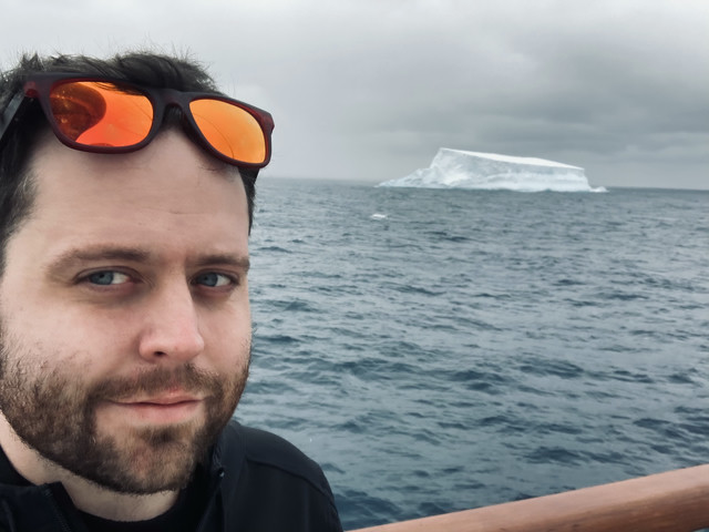 Me taking a photo of myself as we cross the sea to South Georgia. Cold waters behind me, with an iceberg looming in the background.