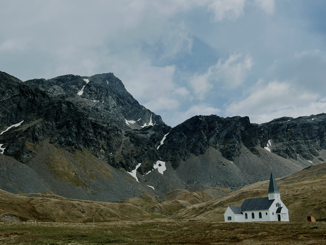 A white church with a dark roof sits in the shadows of a backdrop of cliffs. Green grass and quarry rocks all around the church.