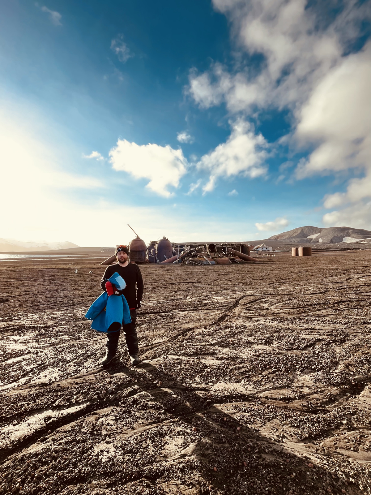 Me dressed in black holding a blue coat, posing in front of some old rusting oil tanks. The ground is silty and soft, and stretches into the distance around me. Two penguins walk nearby. The sky above is blue with clouds.