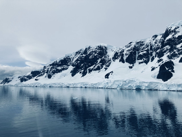 Striking black cliffs with patches of ice and snow line a channel of water. The sky is gray and the reflection of the mountains is visible in the water.