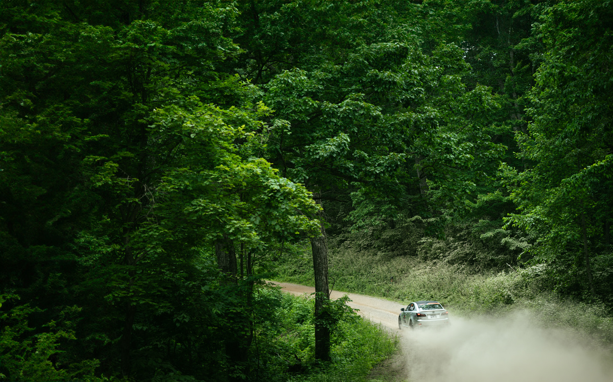 A silver Lexus speeds down a dirt road. Greenery surrounds the road, and dust is being kicked up behind the car.