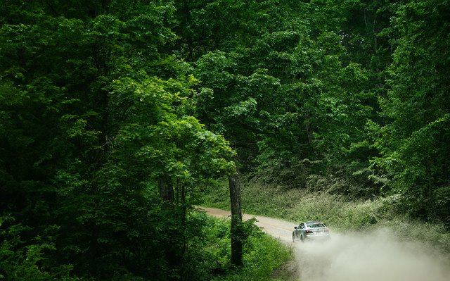A silver Lexus speeds down a dirt road. Greenery surrounds the road, and dust is being kicked up behind the car.