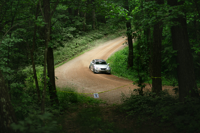 A silver Lexus skims down through a dirt road in a very green lush forest.