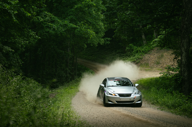 A silver Lexus powers through a dirt road turn, dust kicking up behind it as it heads uphill. The surrounding forest is lush and green.