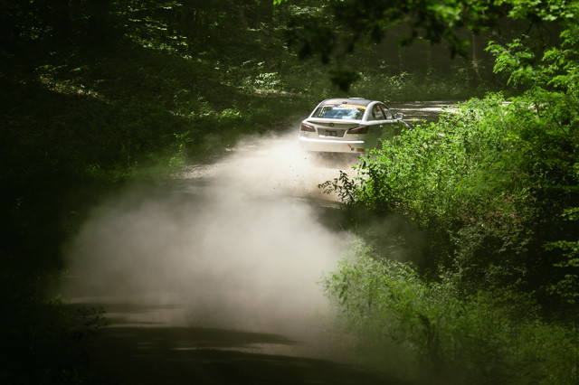 A silver Lexus powers through a long turn in a lush green forest. Dirt clouds are kicked up behind the car.