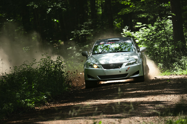 A silver Lexus rips through a turn in the woods. Leaves and dust are kicked up behind it.