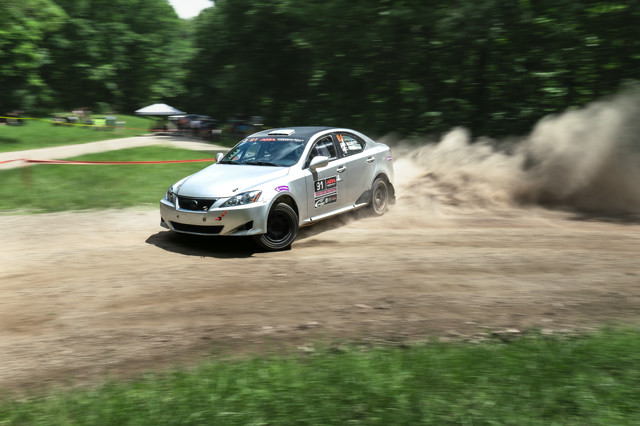 A silver Lexus slides through a wide dirt track turn. A massive dirt cloud is spewed up behind it. Spectator tents are visible in the background.