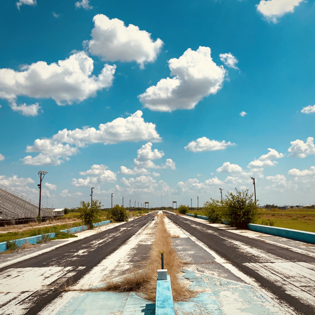 A crumbling drag racing strip stretches in to the distance. The sky above is filled with clouds. Grandstands are visible over on the left side.