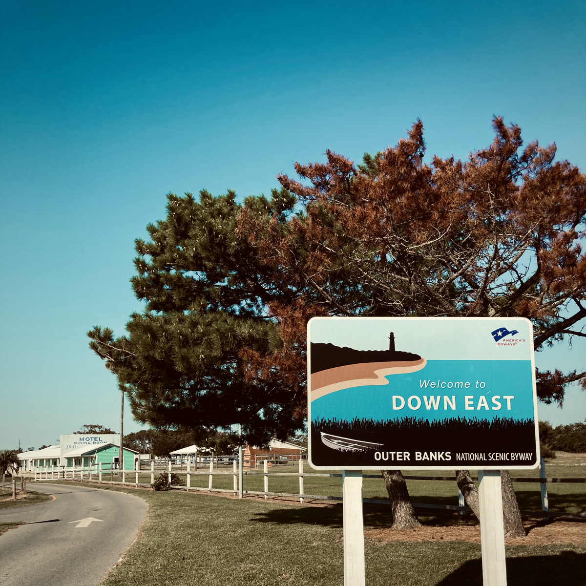 A sign displays text welcoming people to the outer banks. A dilapidated motel is visible in the background.