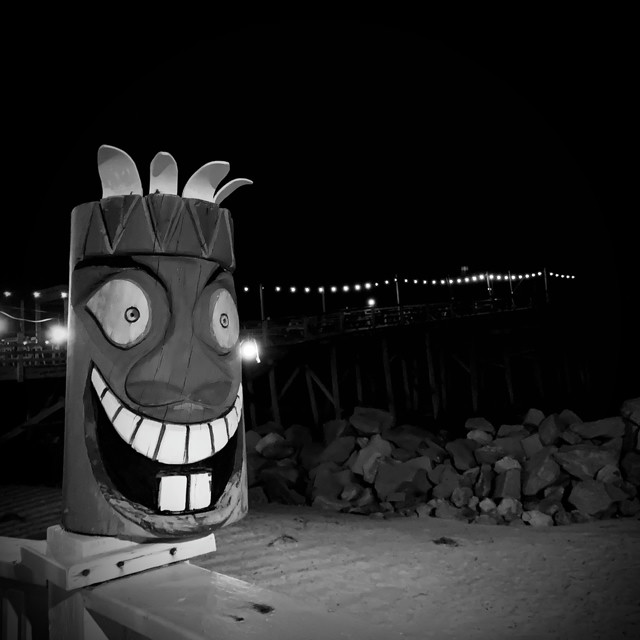 A knockoff crude tiki bar statue is on top of a railing. A pier stretches out over the ocean in the background.