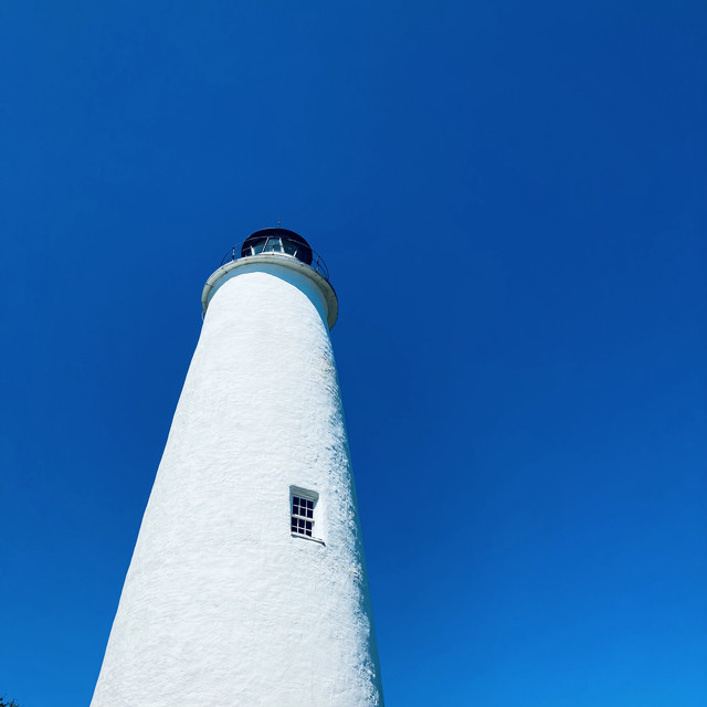 An old white lighthouse rises in to clear blue skies. There’s a window visible on the front.