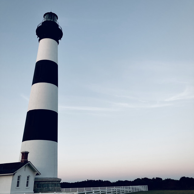 A black and white striped lighthouse at sunset. A white picket fence sits at the bottom.