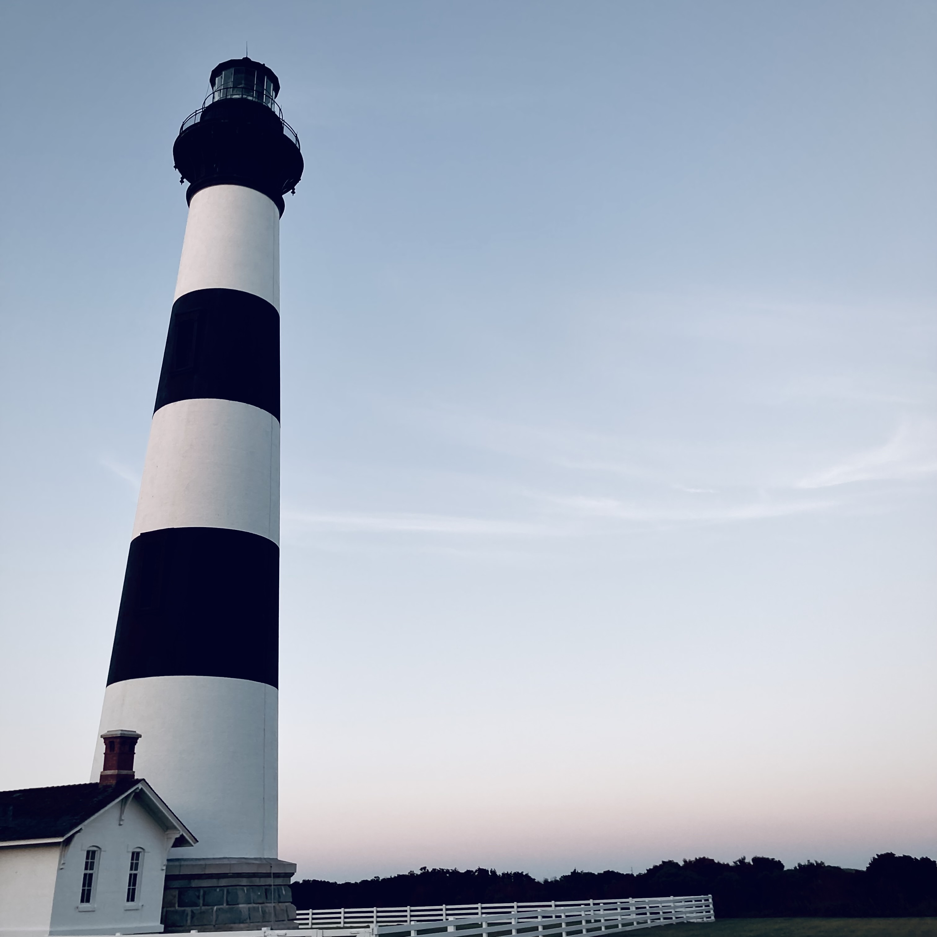 A black and white striped lighthouse at sunset. A white picket fence sits at the bottom.