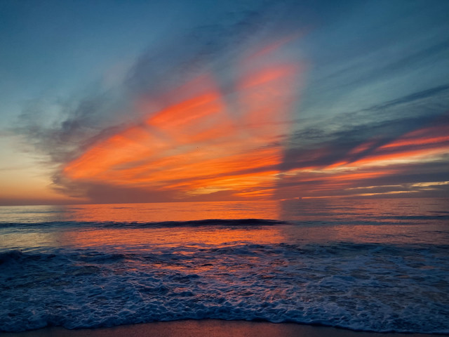 An orange glow floods the far reaches of the ocean. Waves settle on the beach as the clouds split into multiple colors.