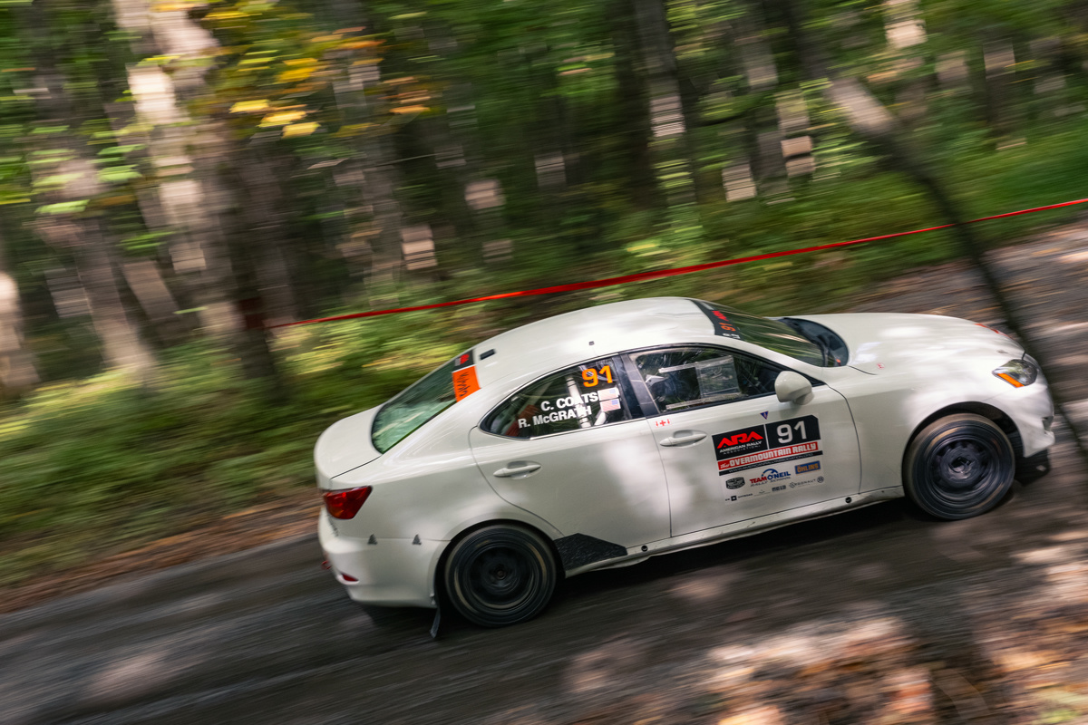 A white rally car racing through the woods at high speed. A red tape barrier is visible in the trees in the background.