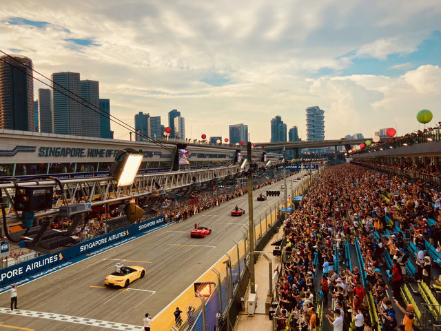 Red and yellow Ferraris are driving down a race track. The grandstands on the right are filled with people. The city looms in the distance.