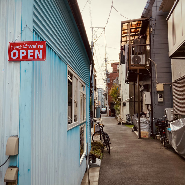 A coffee shop in a blue building in an alleyway in Japan. The alleyway has various bicycles and cars throughout. Laundry can be seen hanging from a balcony as it dries.