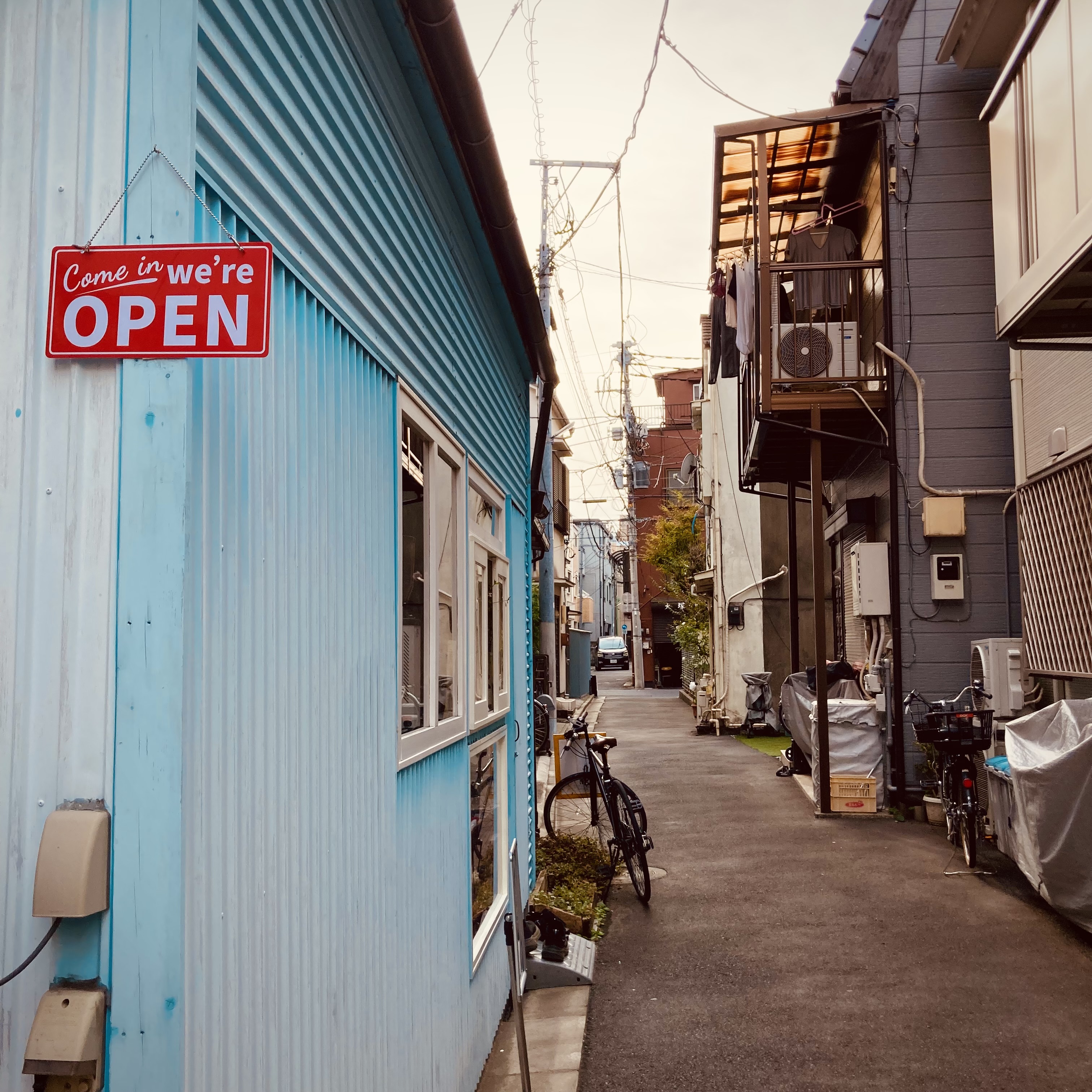 A coffee shop in a blue building in an alleyway in Japan. The alleyway has various bicycles and cars throughout. Laundry can be seen hanging from a balcony as it dries.