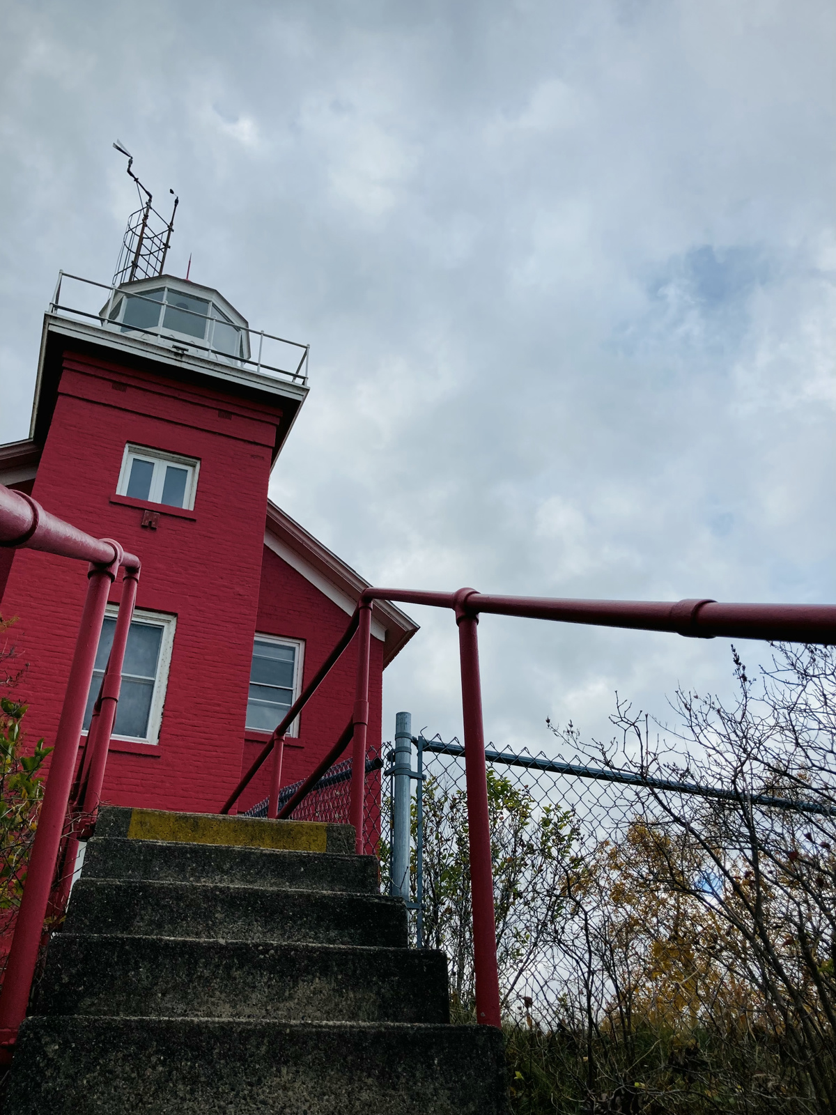 A bright red lighthouse set against a grey sky. Stone steps lead up to it lined by a red banister.