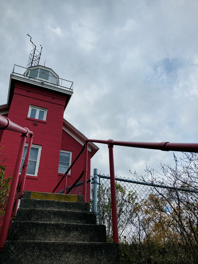A bright red lighthouse set against a grey sky. Stone steps lead up to it lined by a red banister.