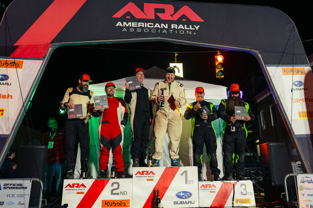 A race podium with spots for 1st, 2nd, and 3rd positions. It's adorned by a large black gate that says 'American Rally Championship'. Sponsor logos can be seen all around. The competitors on the podium are smiling and showing off their trophies.
