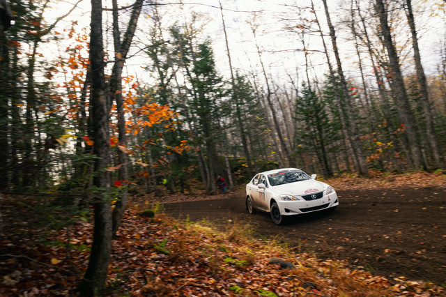 A white Lexus IS250 with dirt on it slides through a dirt turn. The woods behind the car are tight with trees, leaves of endless colors. A cameraman can be seen in the woods behind the car.