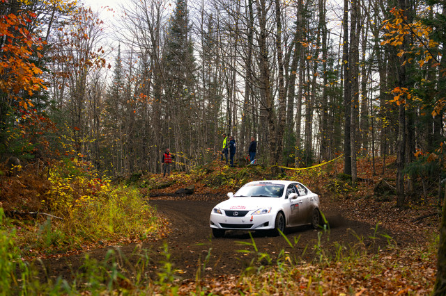 A white Lexus IS250 coming around a right-hand dirt turn. There's a clean swept line, and the Lexus is clearly not on it. People can be seen in between the trees in the background.