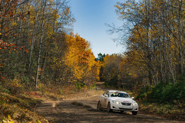 A white Lexus IS250 races through dirt roads. The woods surrounding it are awash in fall colors. Dirt is kicked up from the rear wheels of the car. A photographer can be seen off in the distance.
