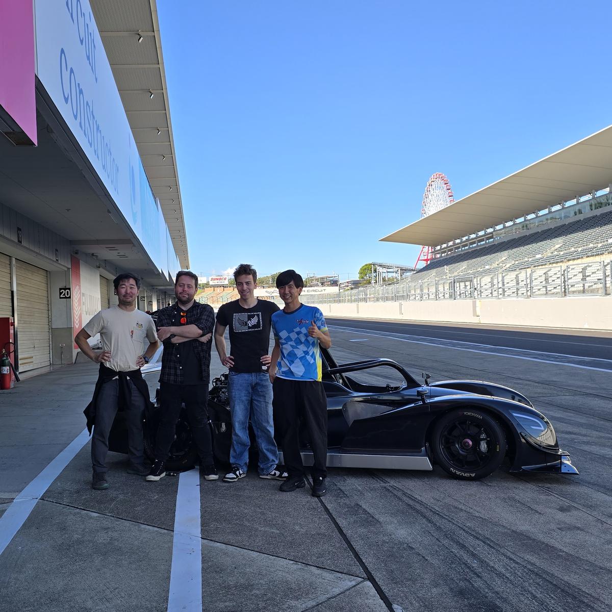 My buddy Tomo, me, and some other crew members standing in front of a black open seater race car. The Suzuka ferris wheel is visible off in the distance.