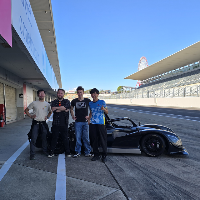 My buddy Tomo, me, and some other crew members standing in front of a black open seater race car. The Suzuka ferris wheel is visible off in the distance.