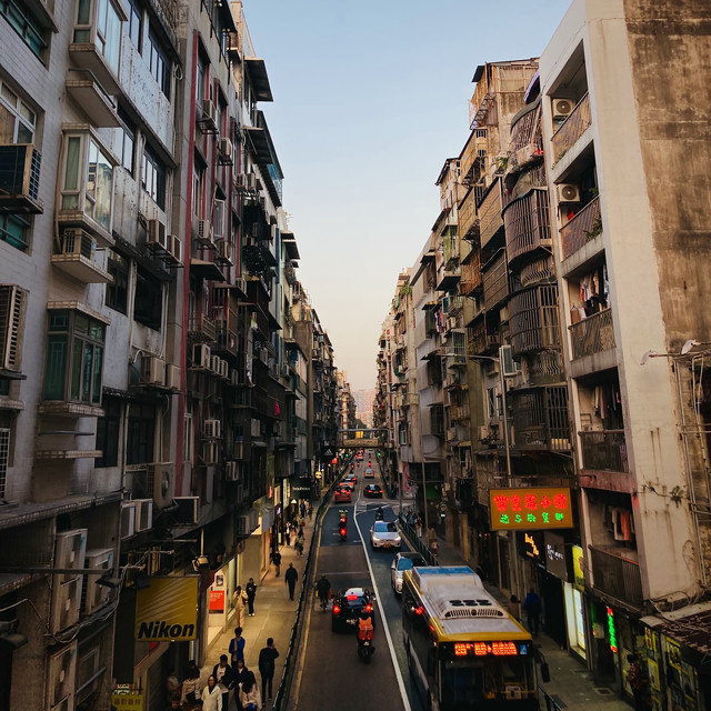 A bustling back street in Macau. The sun is setting overhead. Traffic flows freely and people line the sidewalks as they finish up their day.