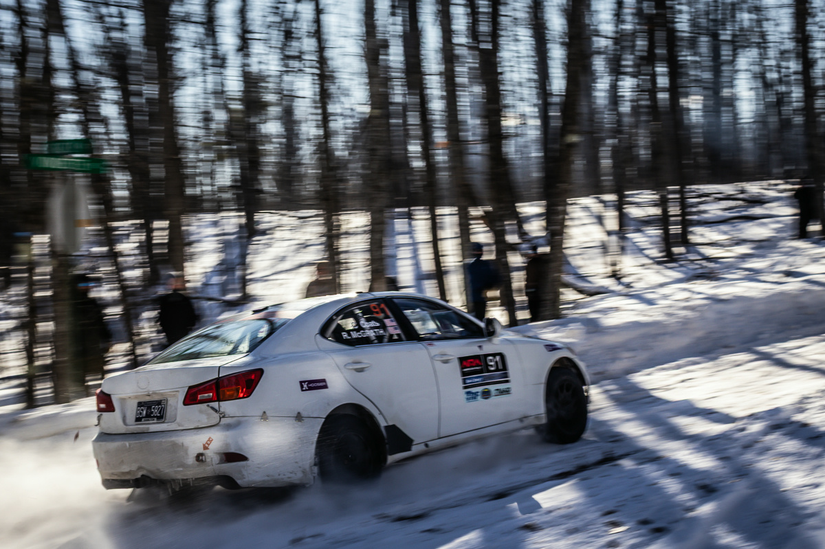 A white Lexus IS250 racing on a snowy icy forest road. Blurred people can be seen in the trees behind it.