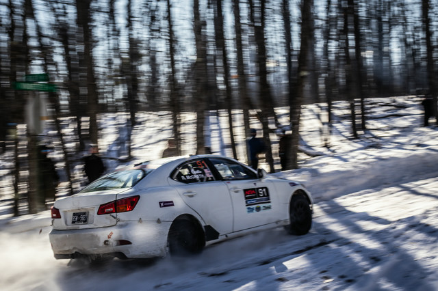 A white Lexus IS250 racing on a snowy icy forest road. Blurred people can be seen in the trees behind it.