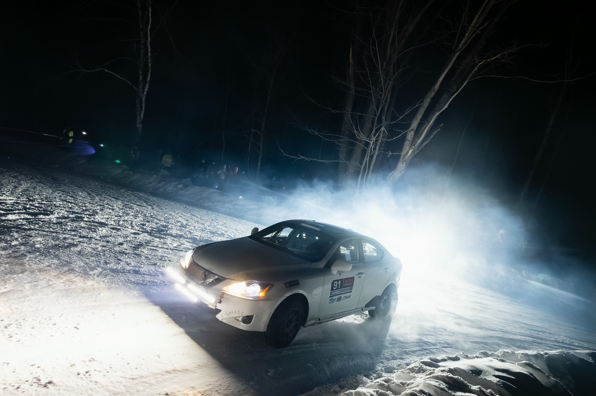 A white Lexus IS250 racing on snowy icy roads. It's mid-turn. Trees line the night sky in the background, with people cheering on from the sidelines.