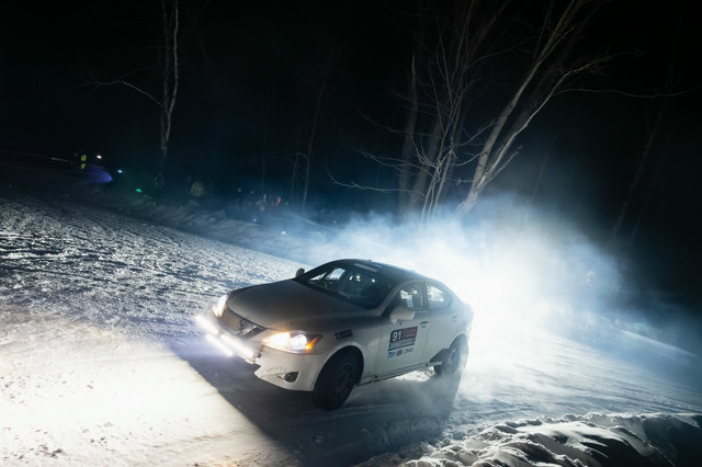 A white Lexus IS250 racing on snowy icy roads. It's mid-turn. Trees line the night sky in the background, with people cheering on from the sidelines.