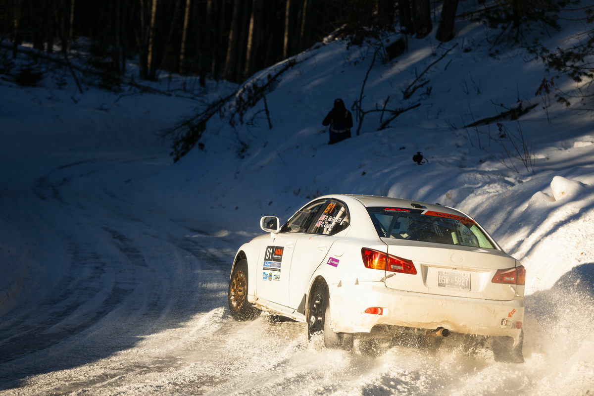A white Lexus IS250 attempts to make a turn on an incredibly icy surface. The sun is setting and casts a ray on the road. The Lexus is sliding due to the ice. A photographer can be seen in the snowy tree-filled background.