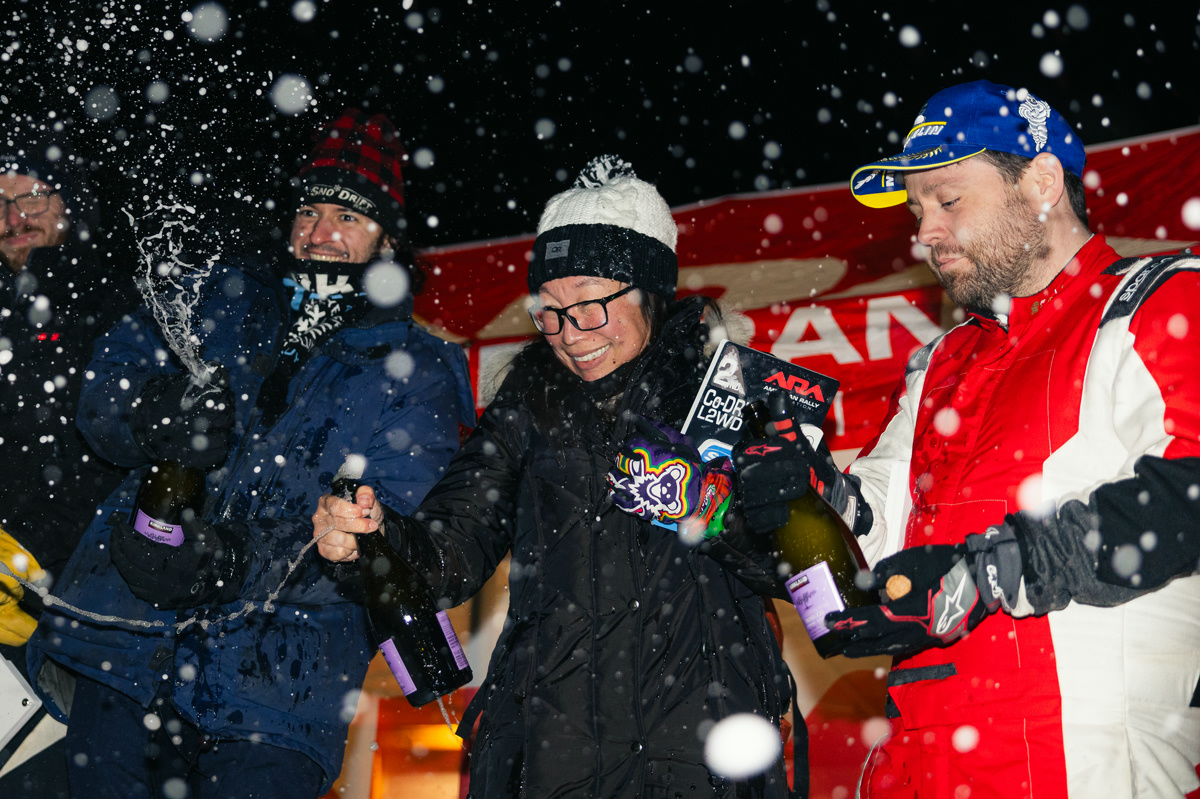 A podium celebration. Multiple contestants are spraying champagne as snowflakes fall.